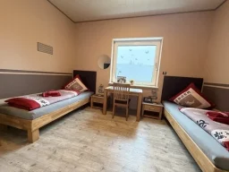 legden-pension-zweibettzimmer-günstig Gemütliches Zimmer in der Pension mit einem Bett, Stuhl, Schreibtisch und kariertem Wanddesign.