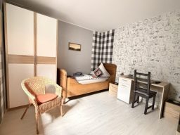 legden-pension-zweibettzimmer-günstig Gemütliches Zimmer in der Pension mit einem Bett, Stuhl, Schreibtisch und kariertem Wanddesign.