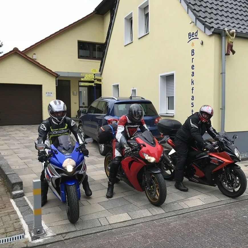 Drei Motorradfahrer auf Sportmotorrädern in der gepflasterten Einfahrt vor der gelben Außenfassade der Pension in Legden.